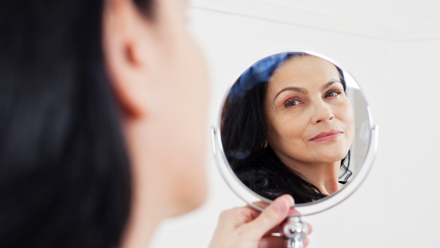 Mature woman with dark hair looking at reflection in hand-held mirror